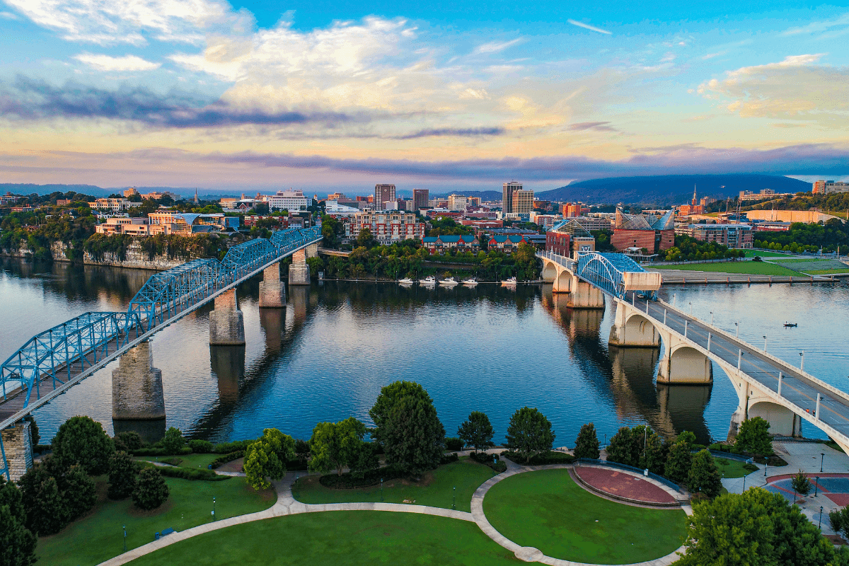 Aerial view of a cityscape featuring two parallel bridges over a river, surrounded by green parks and buildings under a blue sky with scattered clouds.
