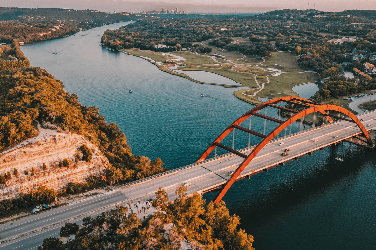 Aerial view of a red arch bridge spanning a wide river, surrounded by lush greenery and hills, with a distant city skyline under a clear sky.