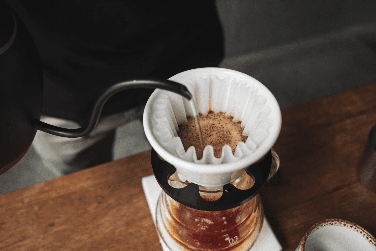 A person is pouring hot water from a kettle into a white ceramic pour-over coffee dripper on a wooden table.