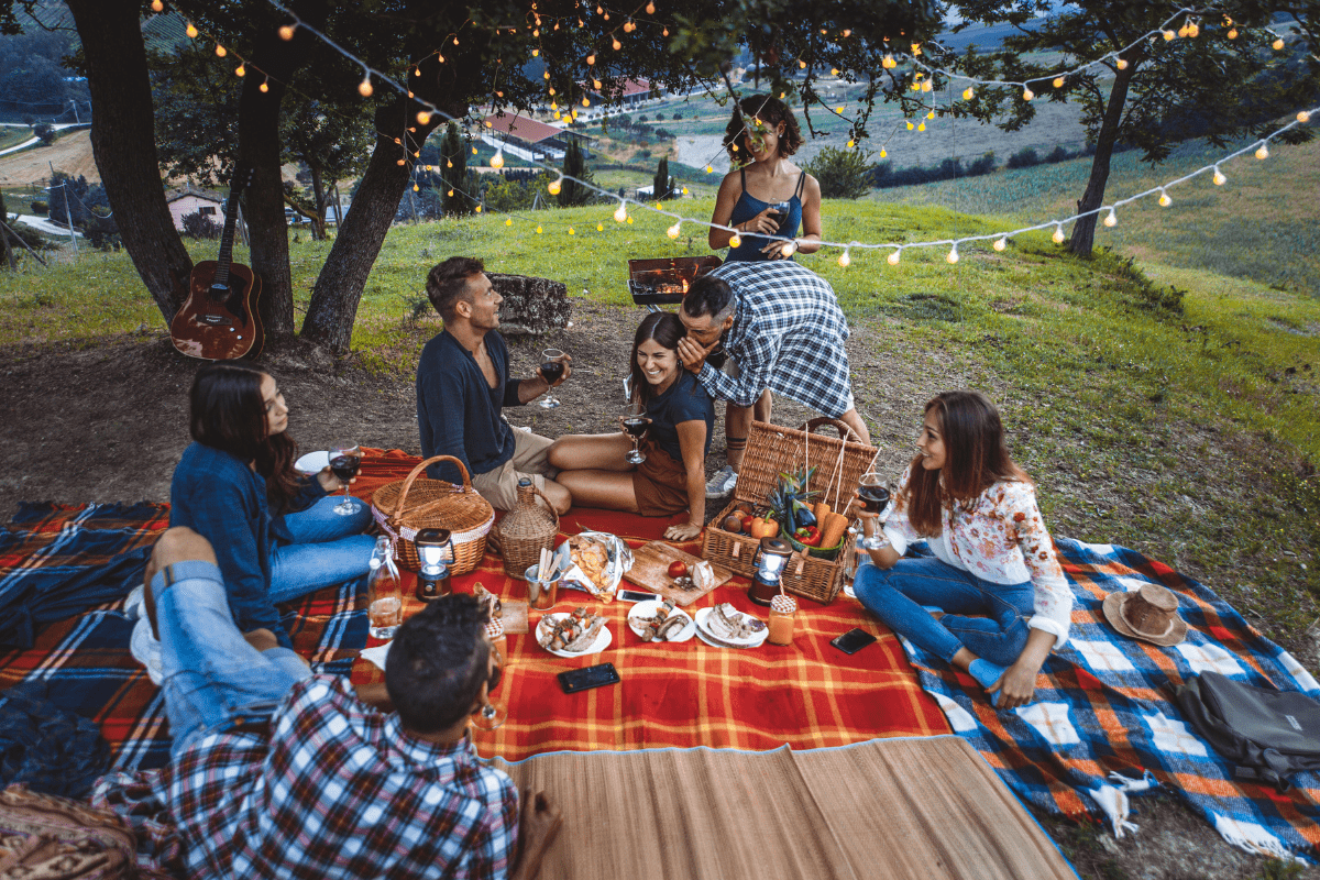 A group of friends enjoys a picnic on colorful blankets under string lights, surrounded by trees, with food, drinks, and a guitar in a scenic outdoor setting.