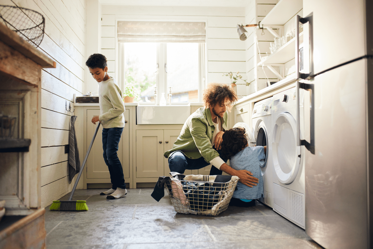 A parent helps a child load a washing machine while another child sweeps the floor in a bright, tidy laundry room.