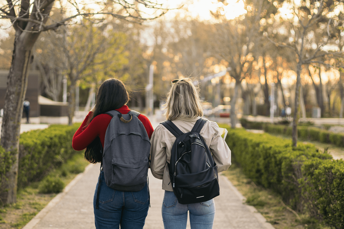 Two people with backpacks walk down a tree-lined path at sunset, surrounded by greenery and soft, golden light filtering through the branches.