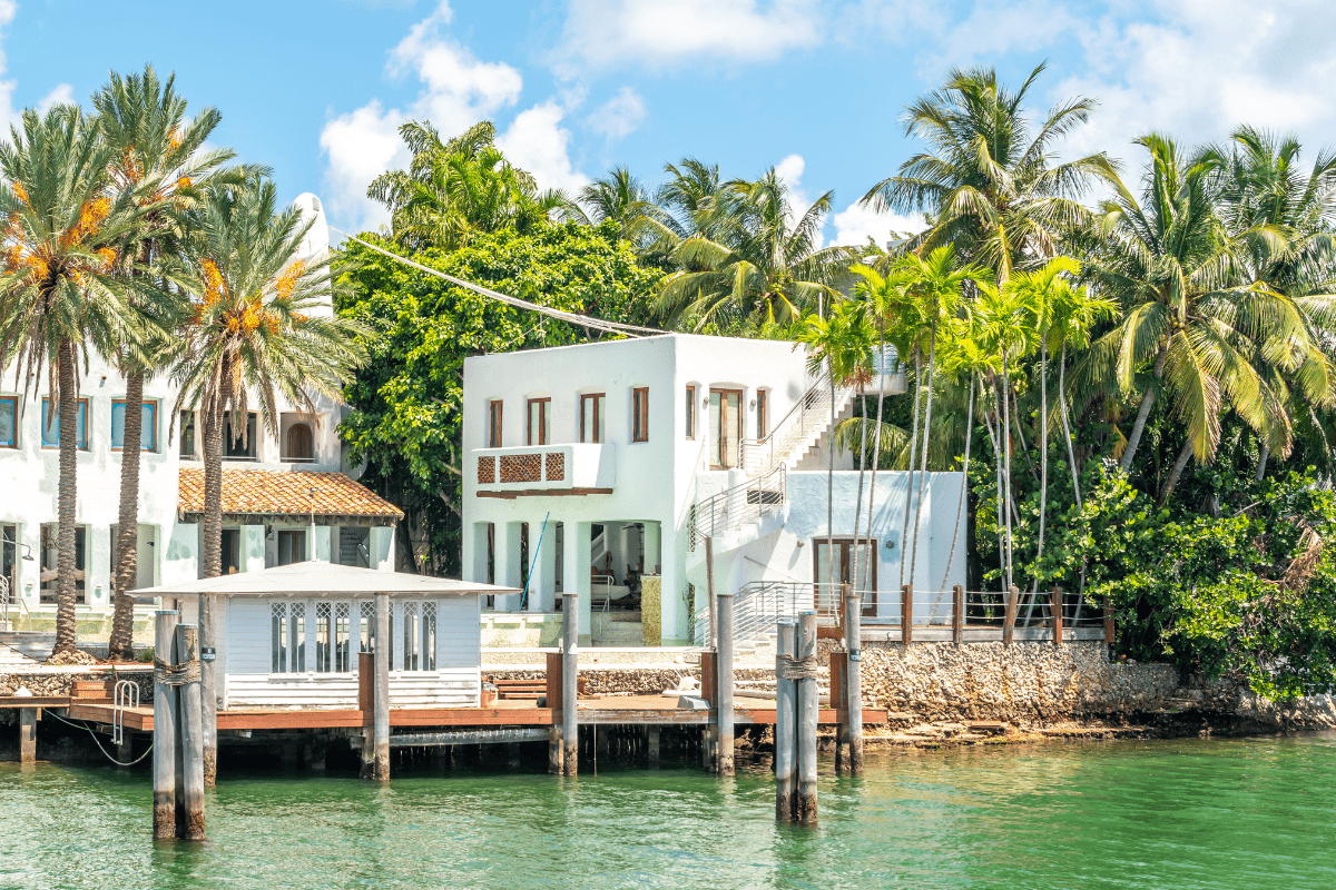 A white, modern waterfront house surrounded by lush palm trees sits beside a wooden dock on a sunny day, with clear blue skies and calm green water.