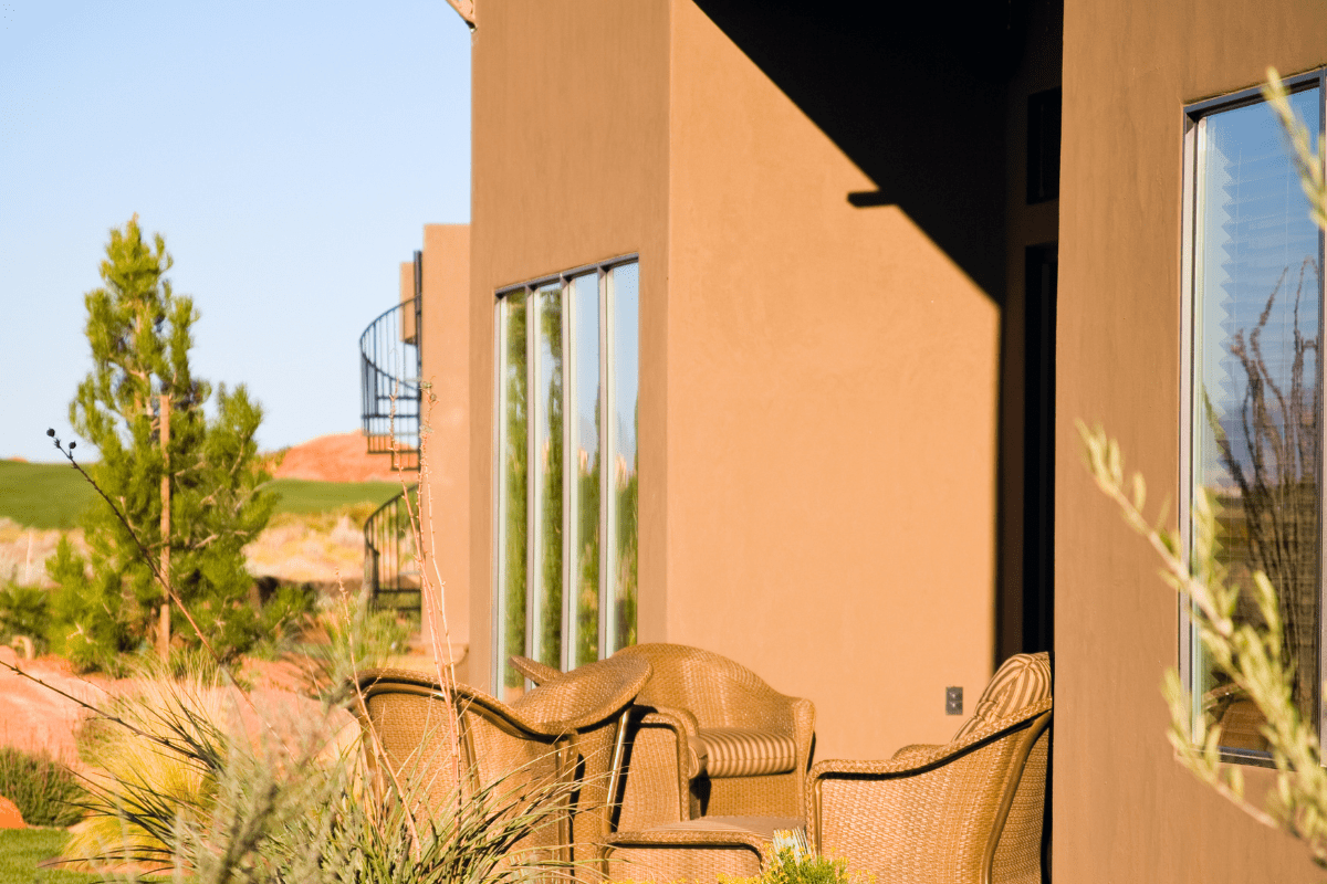 A sunlit patio with wicker chairs is nestled beside a modern brown building, surrounded by desert plants and a spiral staircase in the background.