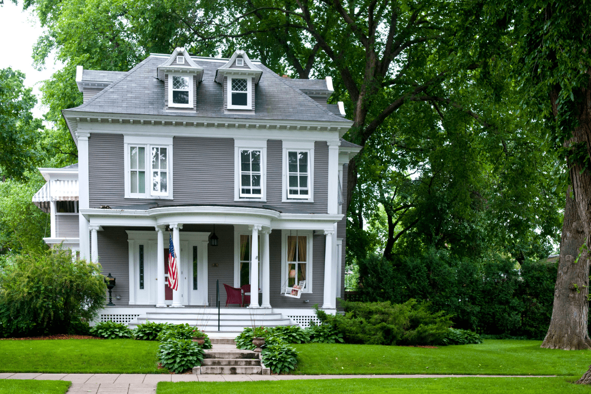 A charming two-story gray house with white trim, featuring a covered porch, American flag, and lush green surroundings, set against a backdrop of tall trees.