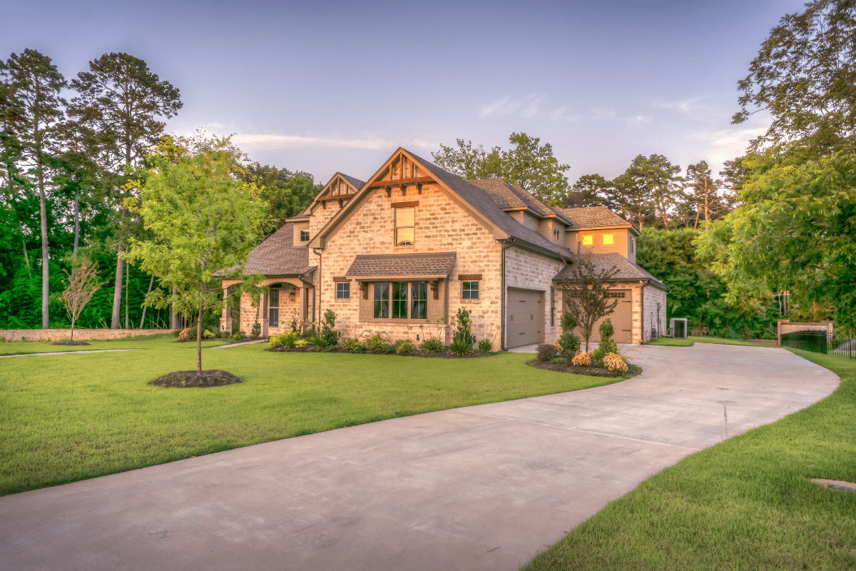 A large stone house with a well-maintained lawn, surrounded by trees, features a curved driveway leading to a two-car garage under a clear sky.