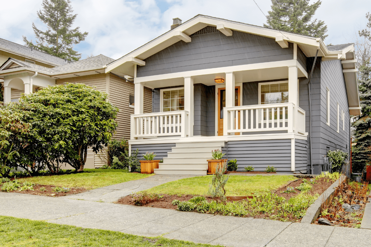 A charming gray bungalow with a small front porch, white trim, and a neatly landscaped yard featuring shrubs and potted plants.