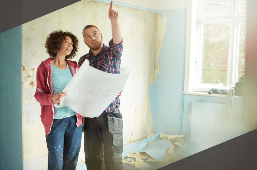 A couple examines blueprints in a partially renovated room, with peeling wallpaper.