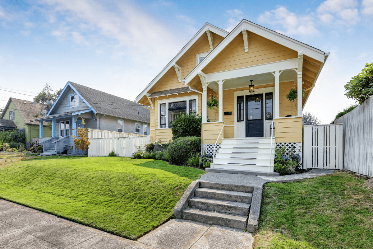 A charming yellow house with a gabled roof, white trim, and a small porch is surrounded by a well-manicured lawn and neighboring homes under a partly cloudy sky.