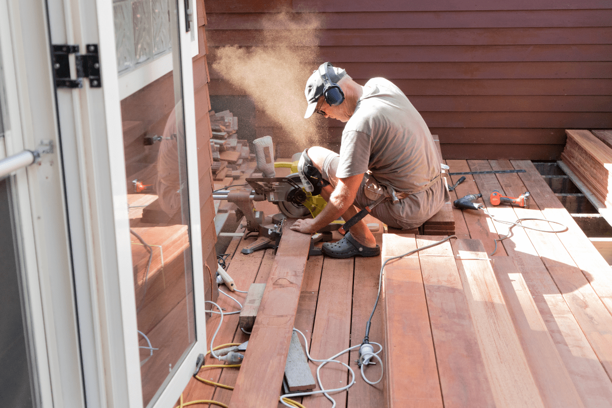 A person wearing protective gear uses a circular saw to cut wooden planks on a deck under construction, surrounded by tools and sawdust.