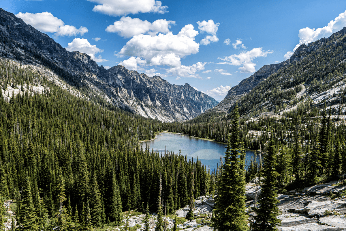 A serene mountain landscape with a clear blue lake surrounded by dense evergreen forests and rugged peaks under a sky dotted with fluffy clouds.