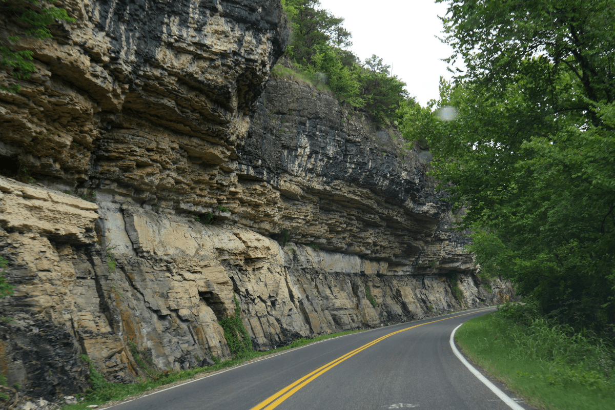 A winding road curves alongside a steep, layered rock cliff, surrounded by lush green trees under an overcast sky.