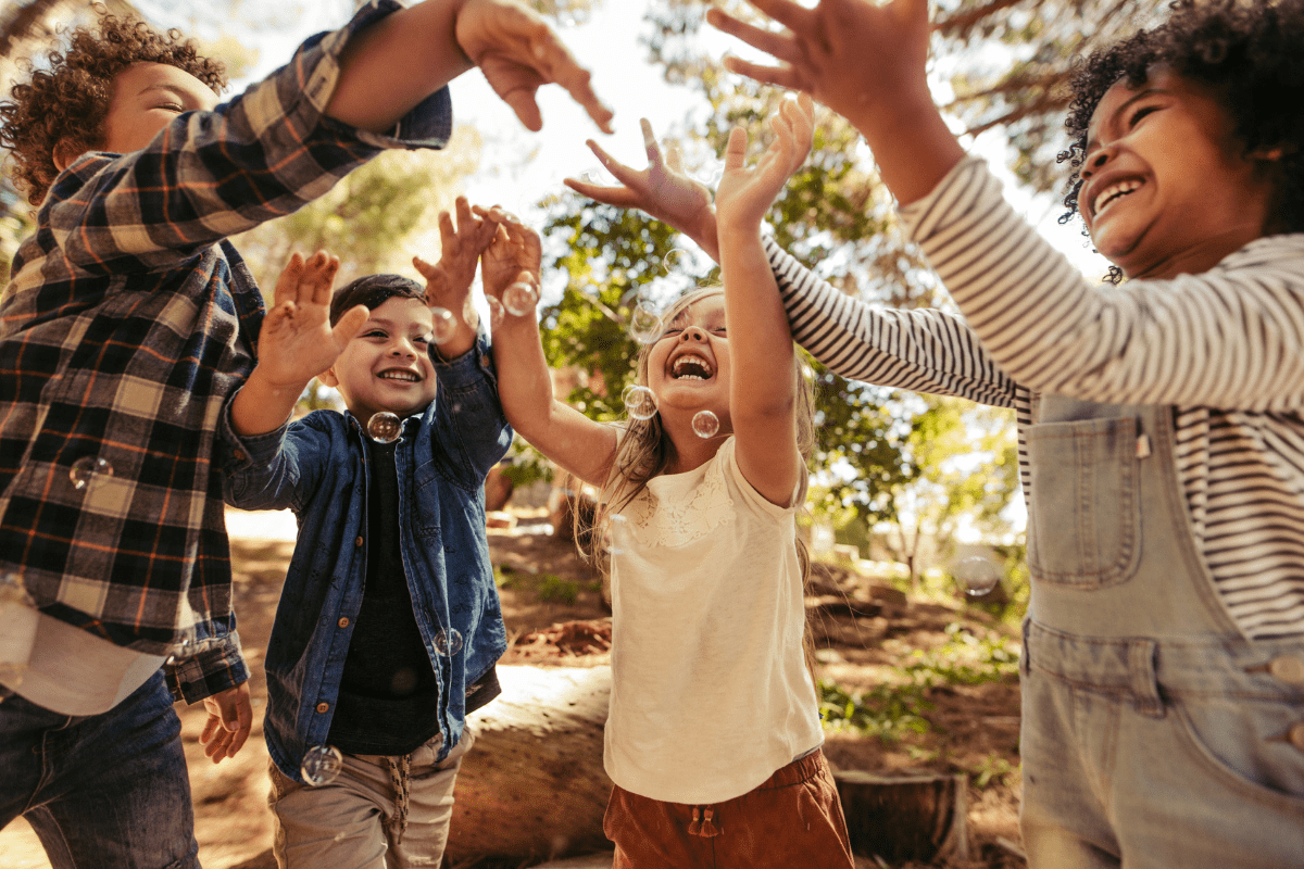 Four children joyfully reach for bubbles in a sunlit park, surrounded by trees, with expressions of delight and excitement on their faces.