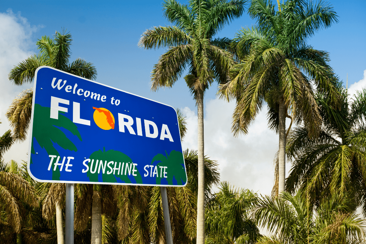 A "Welcome to Florida, The Sunshine State" sign stands against a backdrop of tall palm trees and a partly cloudy blue sky.
