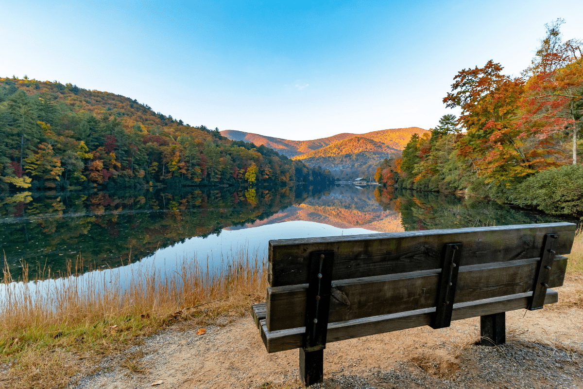 A wooden bench overlooks a tranquil lake surrounded by colorful autumn trees and distant hills under a clear blue sky.