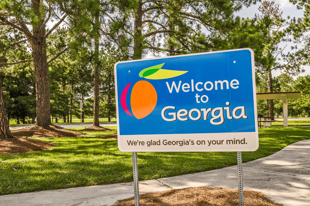 A blue sign with a peach graphic reads "Welcome to Georgia" and "We're glad Georgia's on your mind," set against a backdrop of trees and a grassy area.