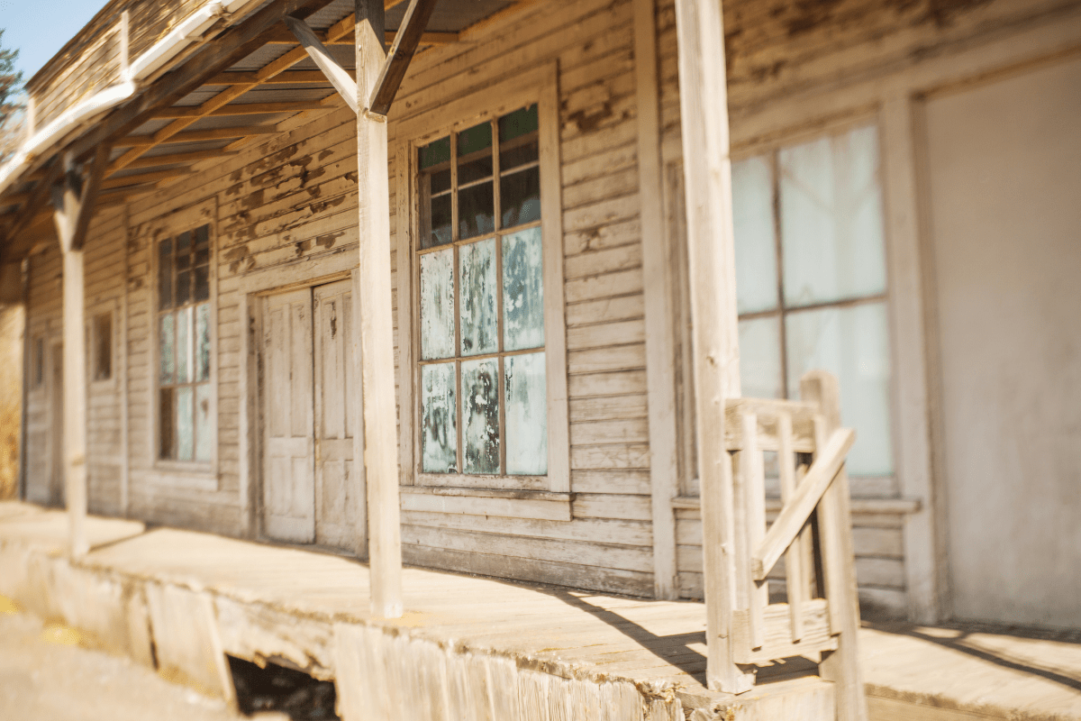 A weathered, wooden building with peeling paint and large, dusty windows stands under a slanted roof, casting shadows on the worn wooden porch in bright sunlight.