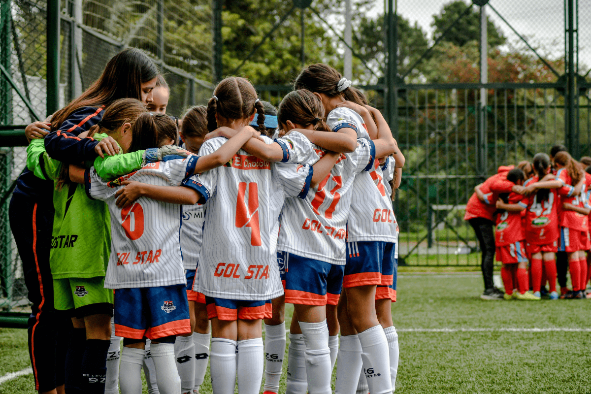 A youth soccer team in white and blue uniforms huddles together on a field, with their coach, while another team in red huddles in the background.