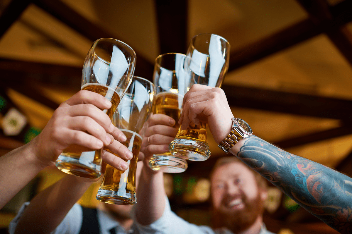 A group of people raise their glasses filled with beer in a celebratory toast, with a focus on their hands and the drinks against a warmly lit background.
