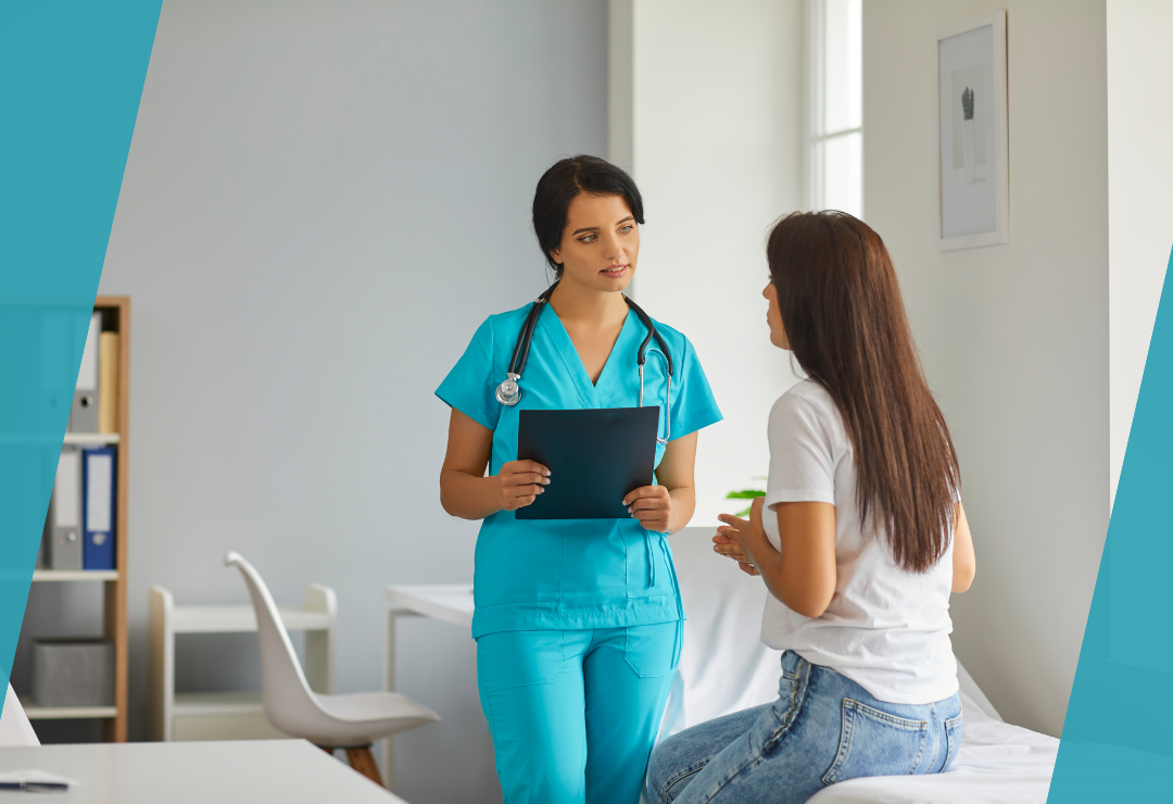 Healthcare professional in blue scrubs talks to a seated patient in a medical office.