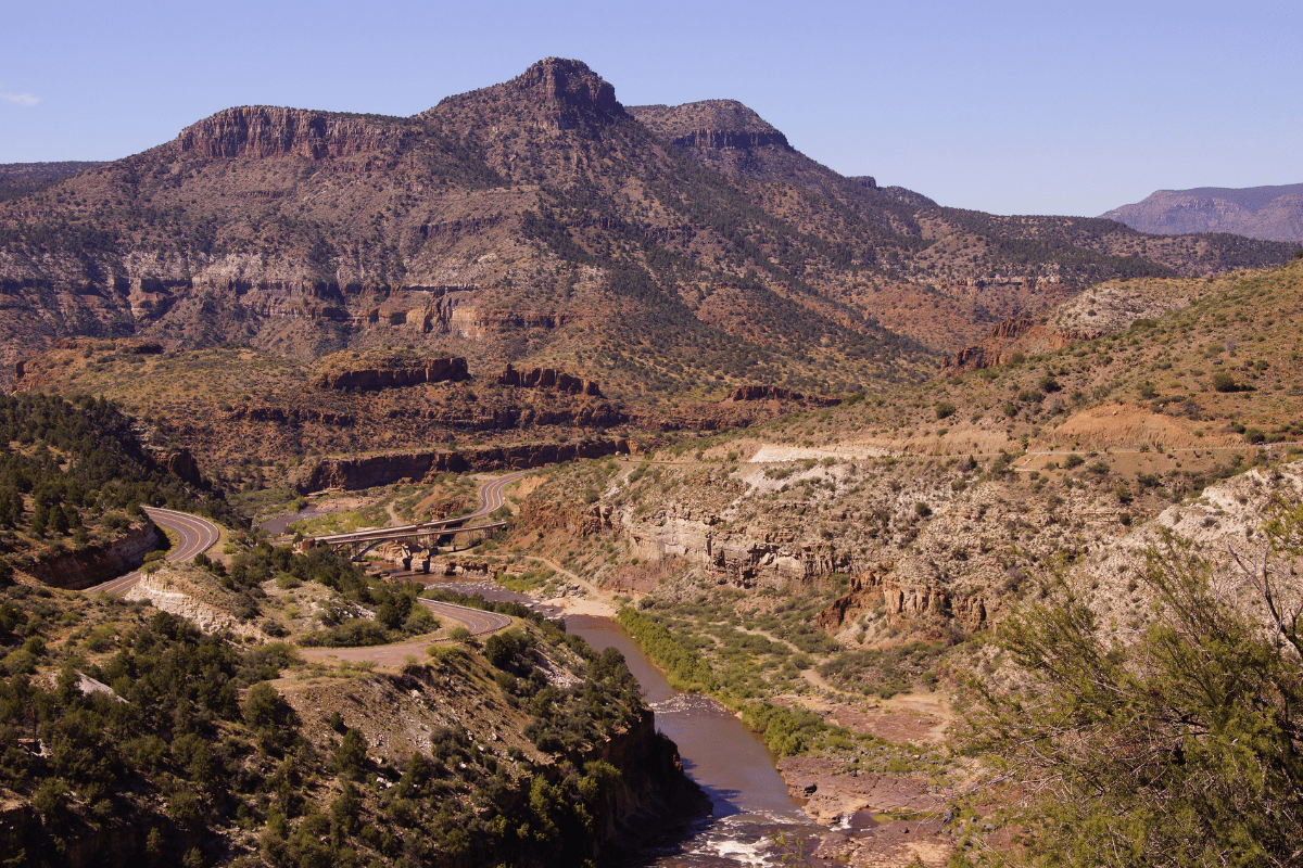 A winding road and river navigate through a rugged, mountainous landscape under a clear blue sky, with sparse vegetation and rocky terrain in the foreground and background.