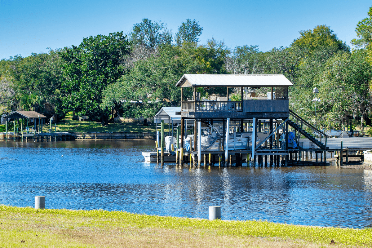A wooden boathouse with a covered upper deck stands over a calm river, surrounded by lush trees and a clear blue sky.