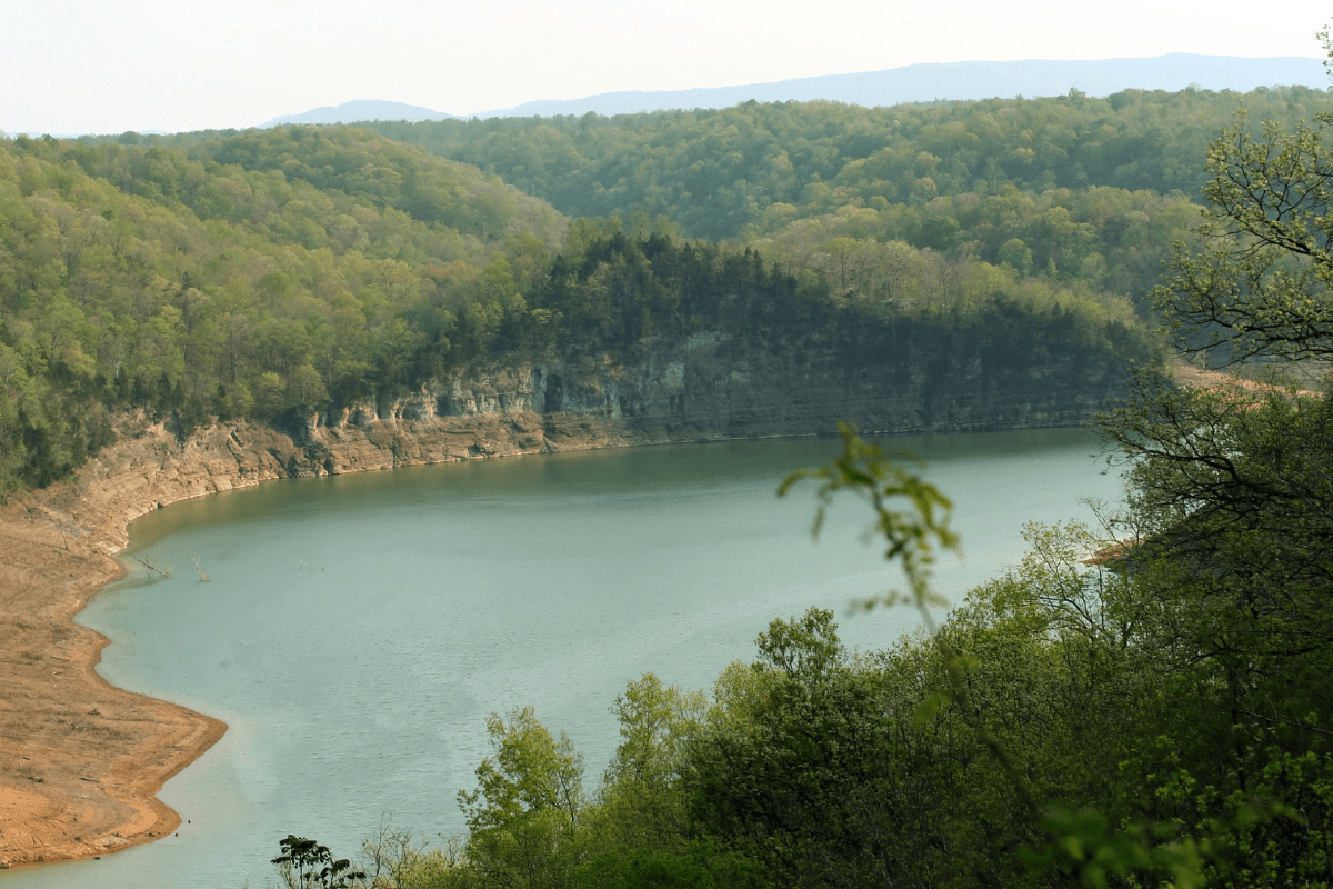 A serene lake is surrounded by lush, green forested hills under a hazy sky, with a rocky shoreline visible in the foreground.