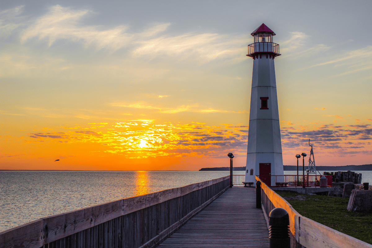 A tall lighthouse stands at the end of a wooden pier, overlooking a calm sea during a vibrant sunset with scattered clouds.