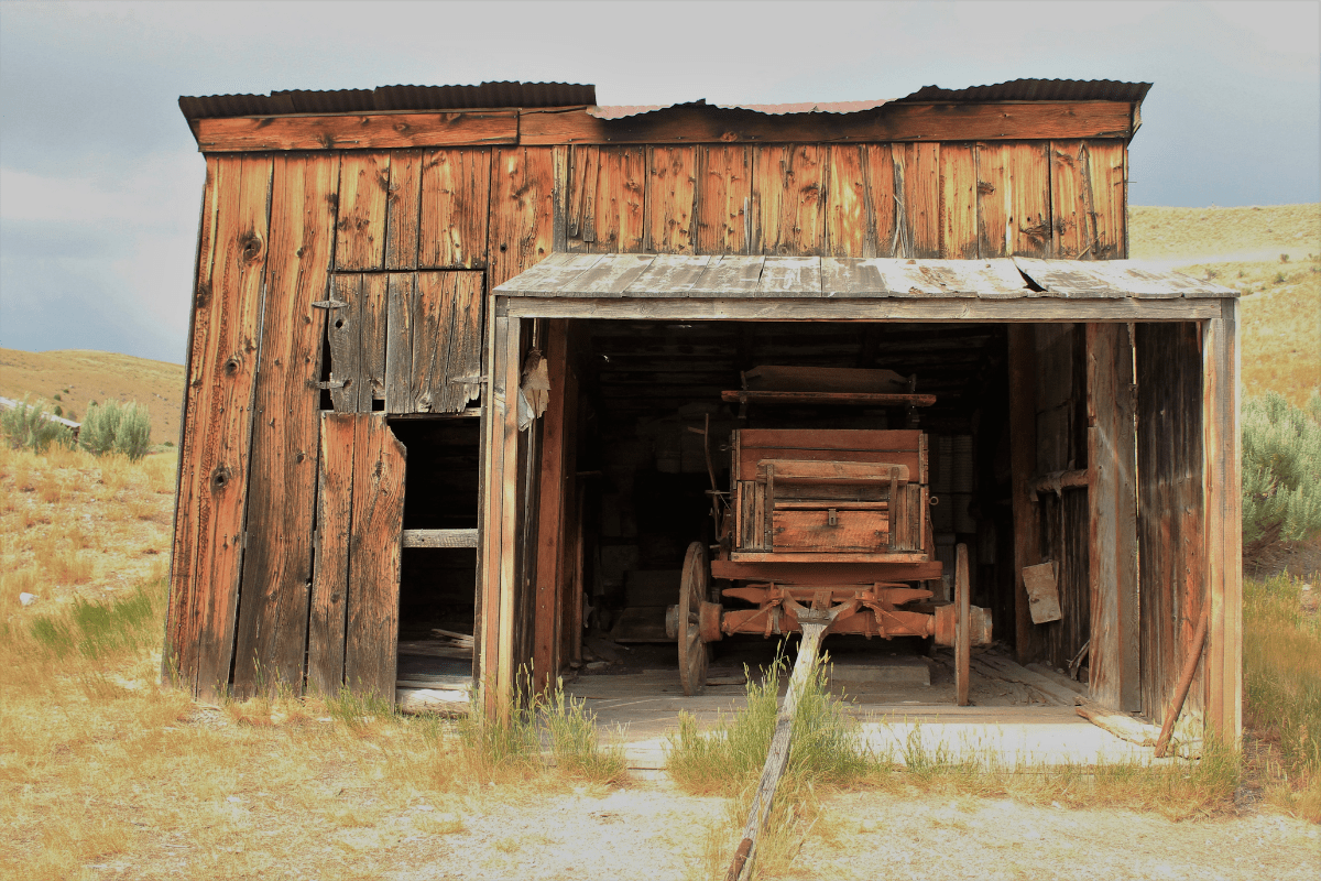 A weathered wooden barn with a partially collapsed roof houses an old, rusted wagon, set against a backdrop of dry grass and a cloudy sky.