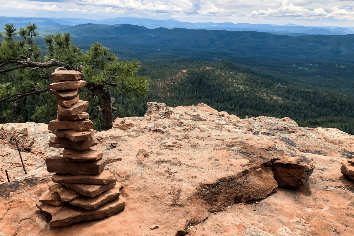 A stack of flat stones balances on a rocky cliff overlooking a vast, forested landscape with distant mountains under a partly cloudy sky.