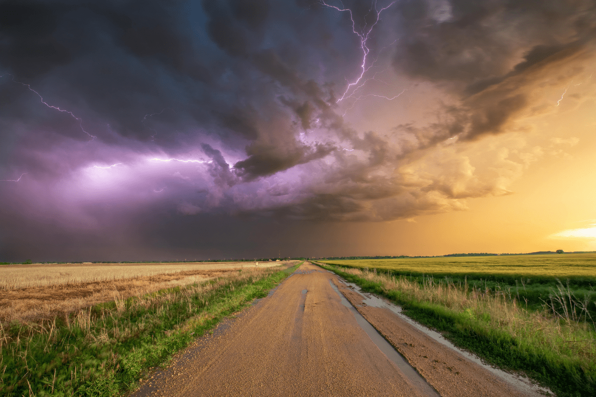 A rural road stretches into the distance beneath a dramatic sky, with dark storm clouds and vivid purple lightning on the left, contrasting with a golden sunset on the right.