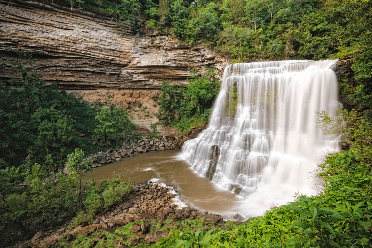 A wide waterfall cascades over layered rock formations into a calm pool, surrounded by lush green foliage and rugged cliffs.