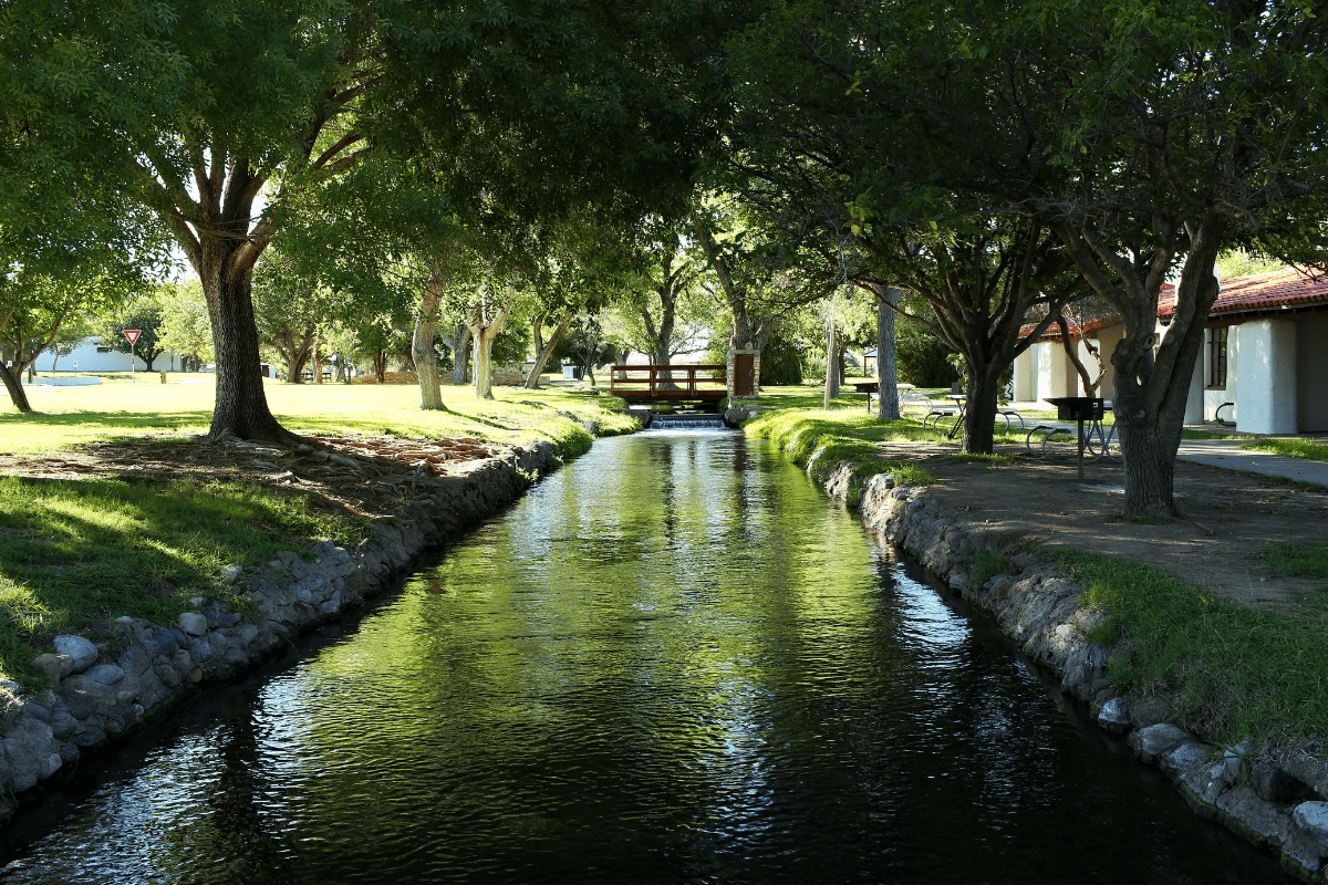 A tranquil stream flows under a wooden bridge, surrounded by lush green trees and a small building with a red roof on the right.