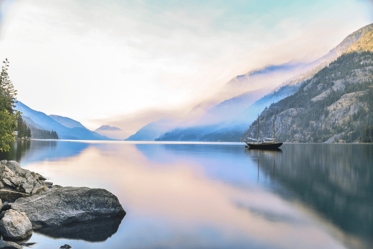A serene lake reflects the soft hues of a pastel sky, with a sailboat anchored near rocky shores and misty mountains in the background.
