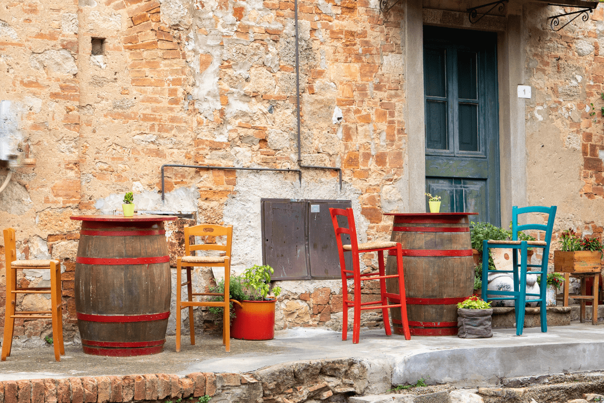 Outdoor seating with colorful wooden chairs and tables made from barrels, set against a rustic brick wall with potted plants.