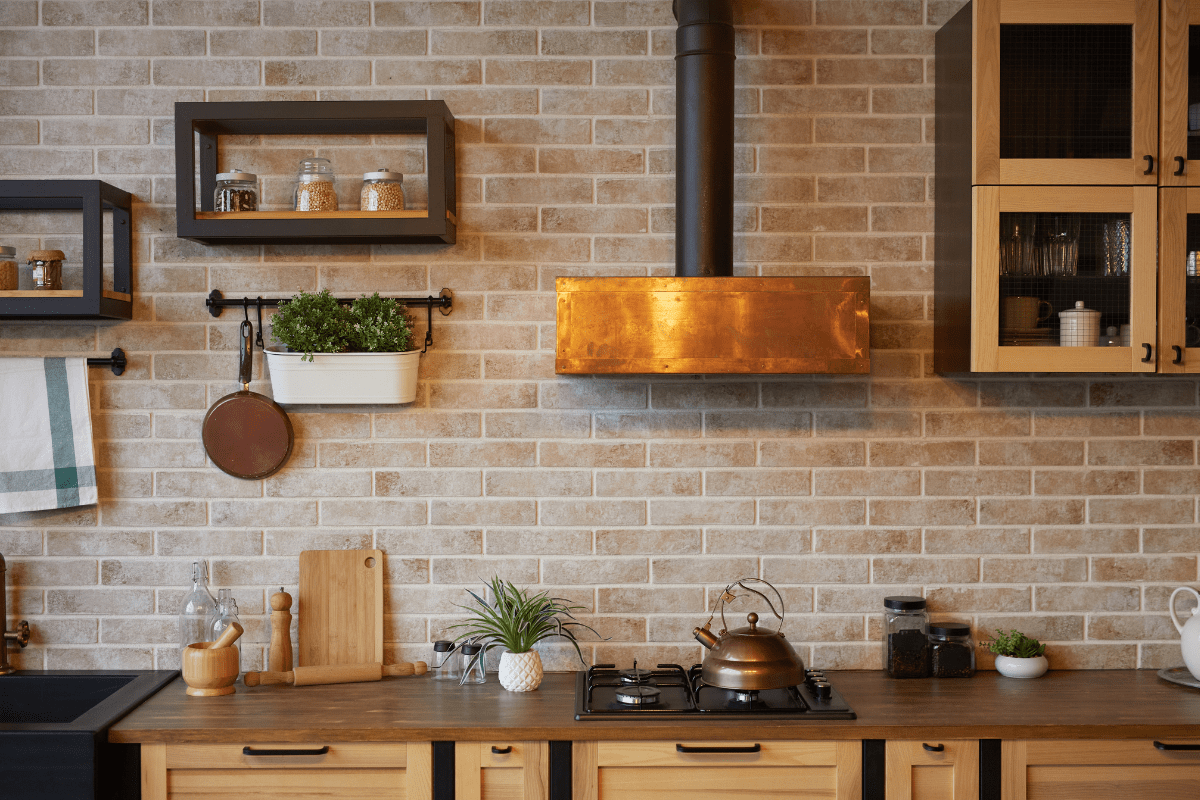 A modern kitchen features a brick backsplash, wooden cabinets, a copper range hood, and a countertop with a kettle, cutting board, plants, and jars.