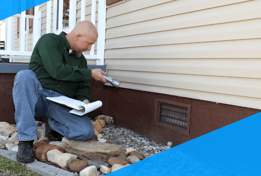 Man inspecting house foundation with flashlight, holding clipboard, surrounded by rocks and stones.