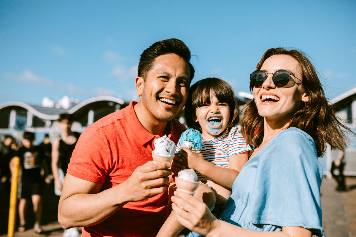 A smiling family of three enjoys ice cream cones outdoors on a sunny day, with the child playfully showing a blue-stained mouth.