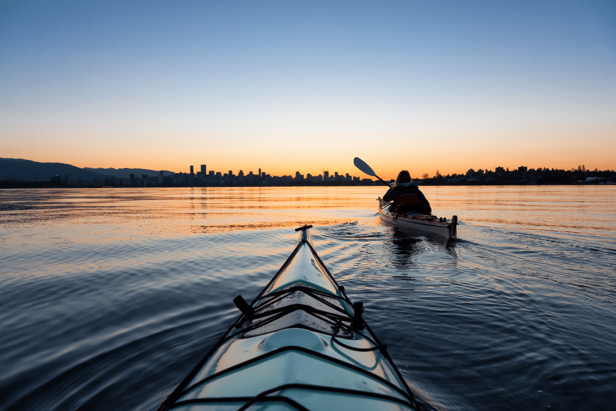 A person paddles a kayak on calm water at sunset, with a city skyline silhouetted against the colorful sky in the background.