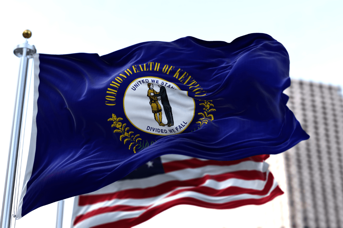 The image shows the Kentucky state flag waving in the wind, featuring the state seal with two figures and the motto "United We Stand, Divided We Fall," against a blurred background of another flag.