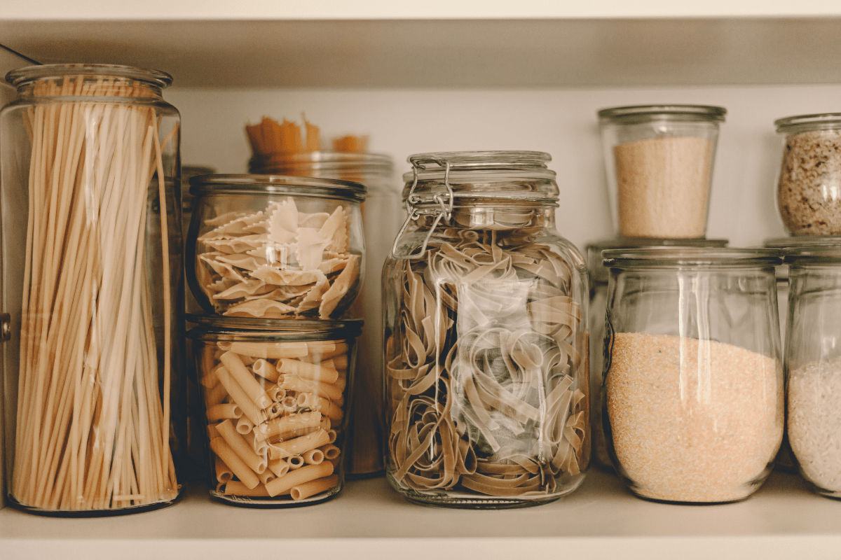 A pantry shelf with glass jars containing various types of pasta, rubber bands, and grains, neatly organized and visible through the transparent containers.