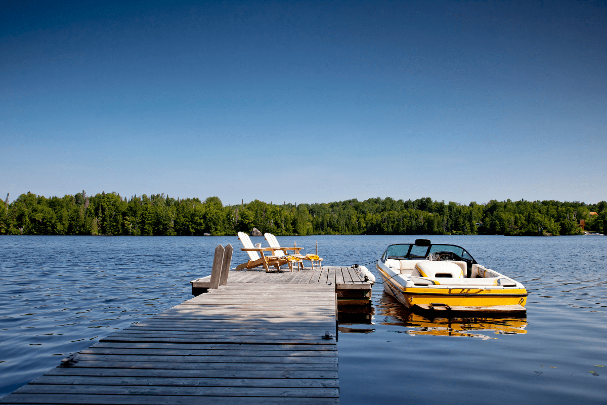 A wooden dock extends over a calm lake with two chairs and a yellow speedboat, surrounded by lush green trees under a clear blue sky.