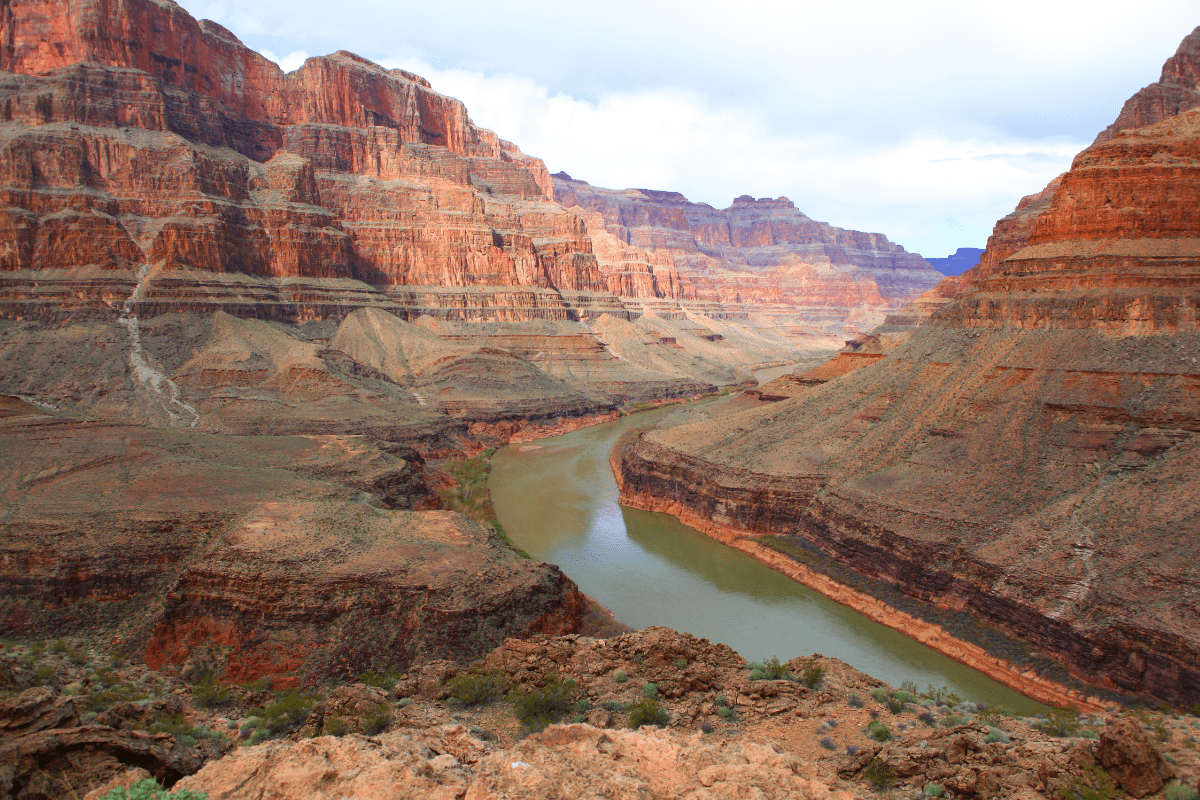 A vast canyon landscape with layered red rock formations and a winding river flowing through the valley under a partly cloudy sky.