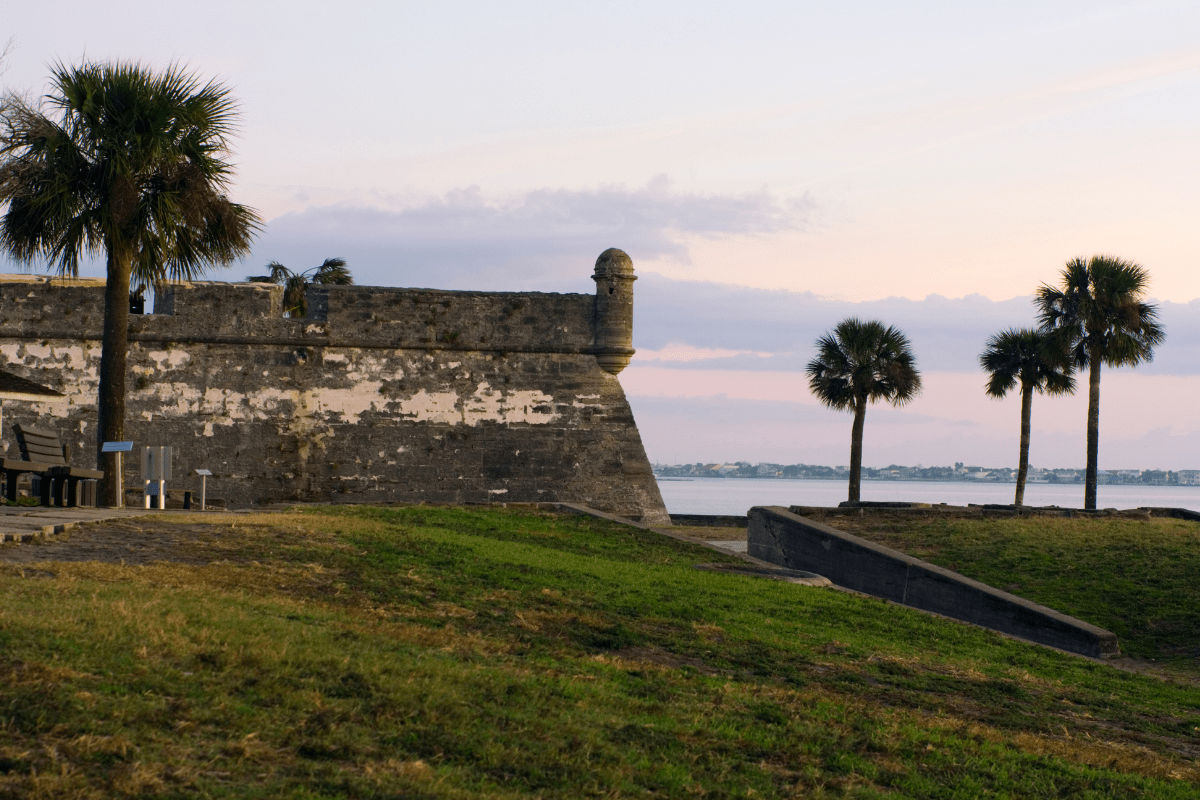Historic stone fort with palm trees in the foreground, set against a serene waterfront and pastel sky at sunset.