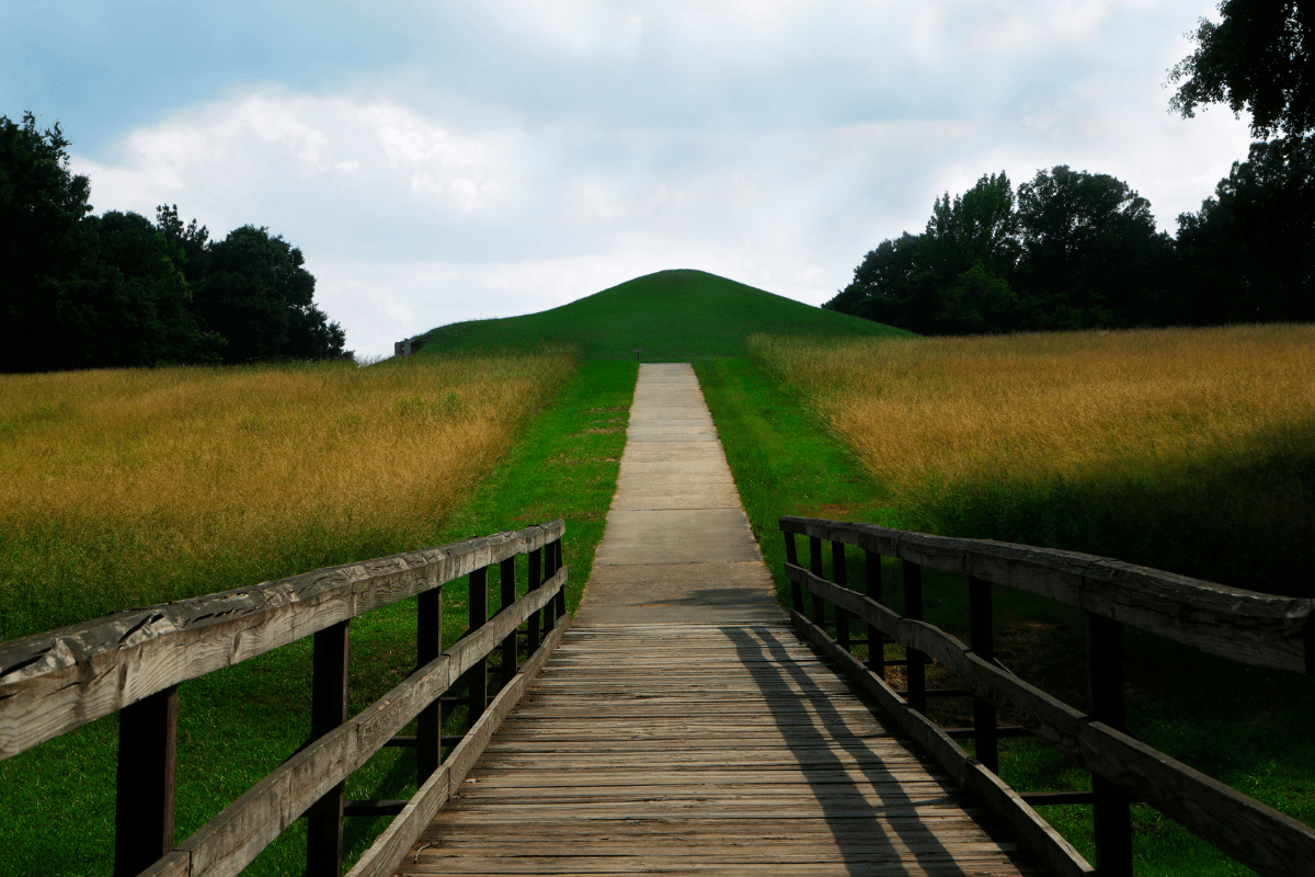 A wooden bridge leads to a paved path through grassy fields, culminating at a large, green mound under a partly cloudy sky.