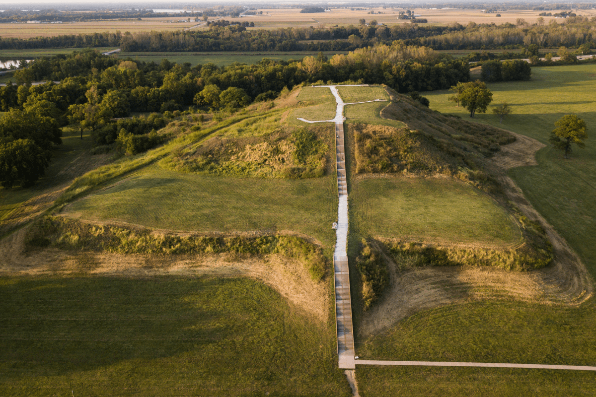 Aerial view of a large, grassy mound with a wooden walkway leading to the top, surrounded by flat, open fields and distant trees.