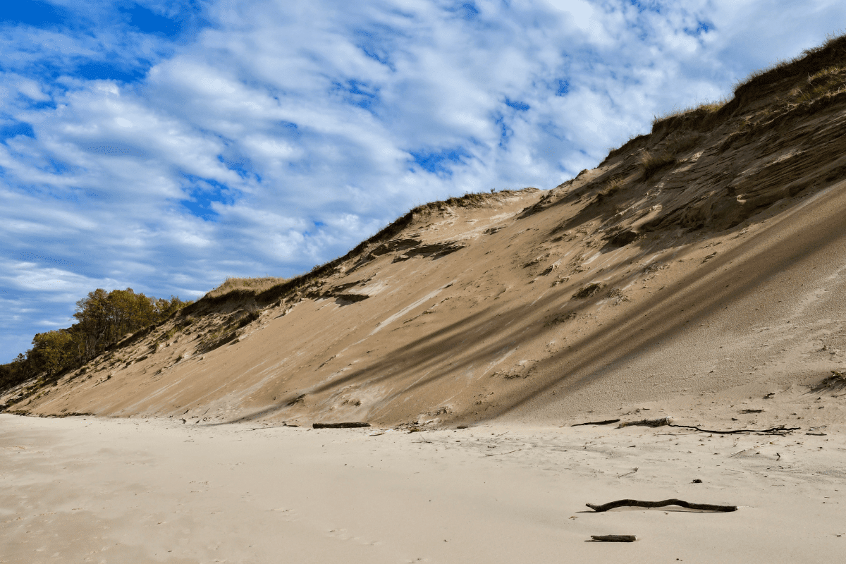 A steep sand dune under a partly cloudy sky, with sparse vegetation on top and driftwood scattered on the sandy beach below.