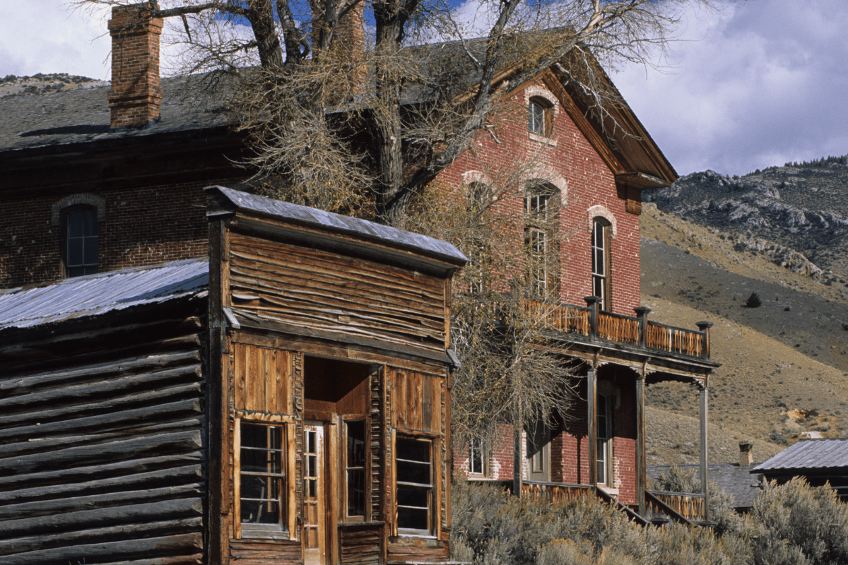 A rustic wooden cabin and a large brick house stand in a mountainous landscape, with leafless trees and a cloudy sky in the background.