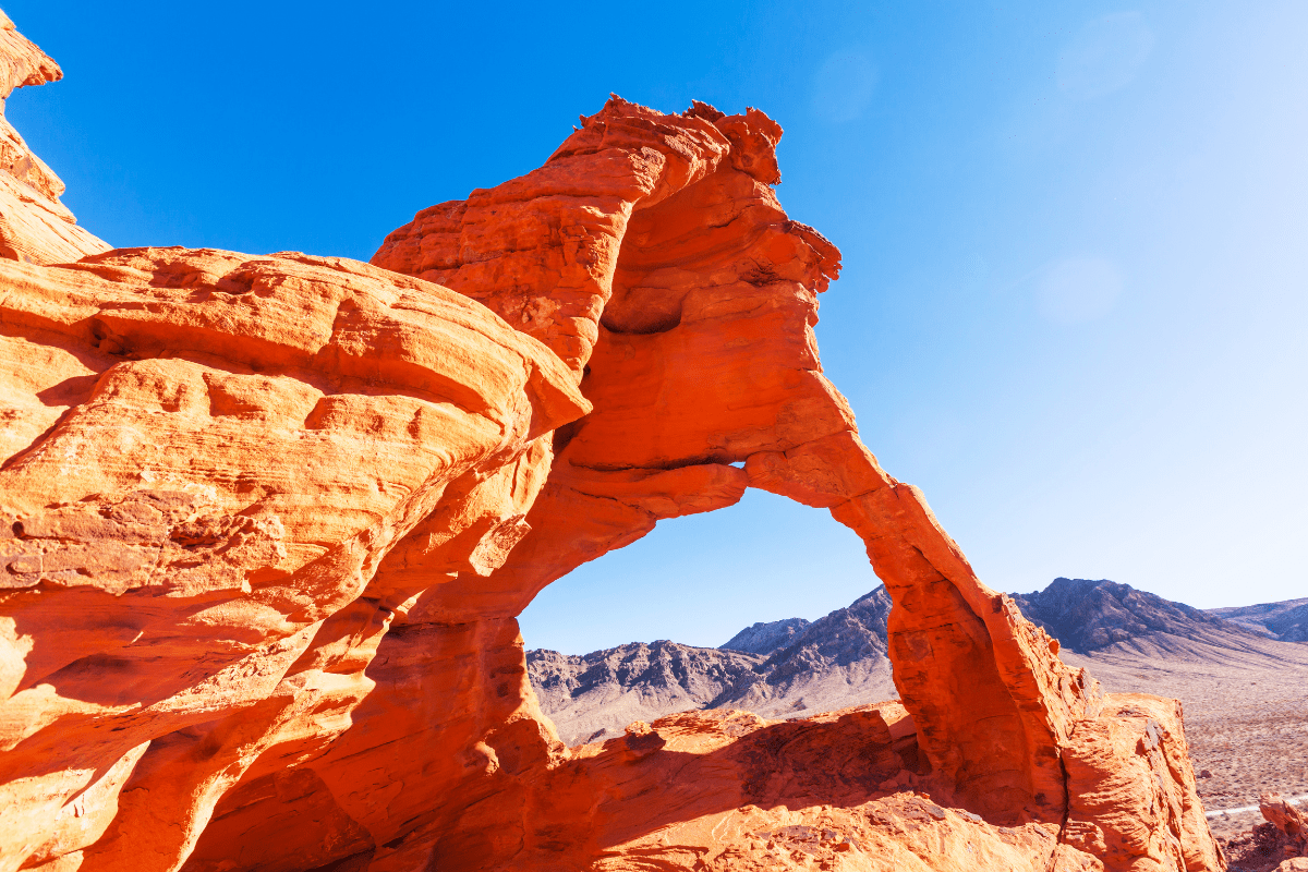 A vibrant red rock arch stands against a clear blue sky, with rugged mountains in the background, capturing the striking desert landscape.