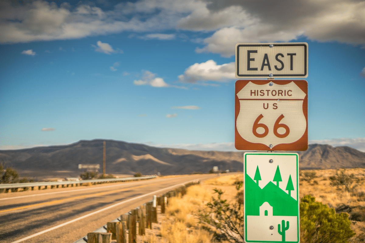 A road sign for Historic Route 66 heading east stands beside a desert highway, with distant mountains and a partly cloudy sky in the background.